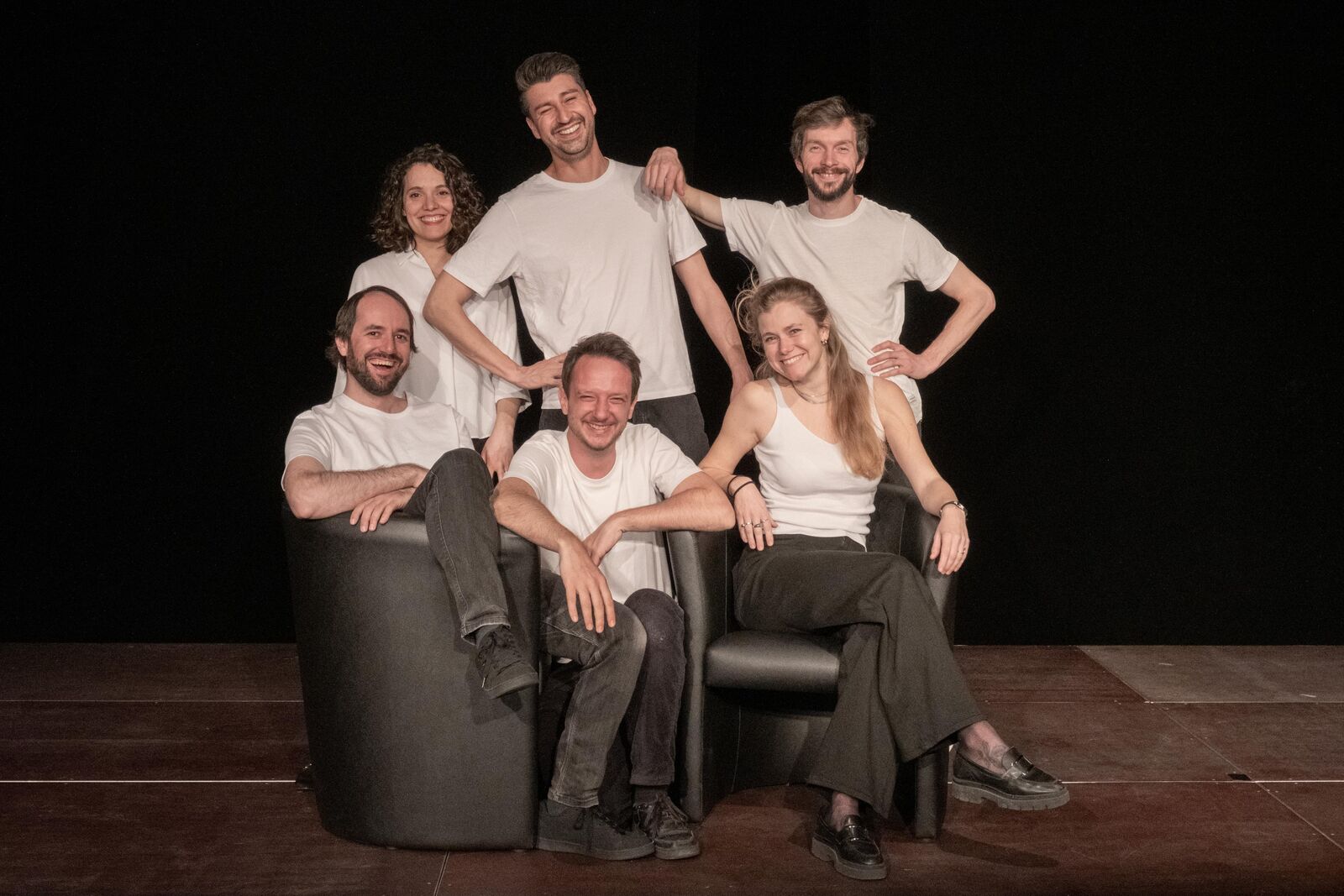 Die Gruppe auf der Bühne: sechs Personen in weissen Oberteilen, auf und hinter zwei Sesseln, vor schwarzem Vorhang.